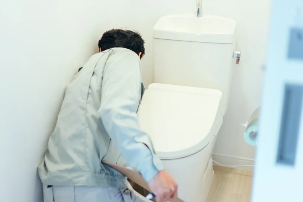 A person in work clothes inspecting or fixing a white toilet in a bathroom.