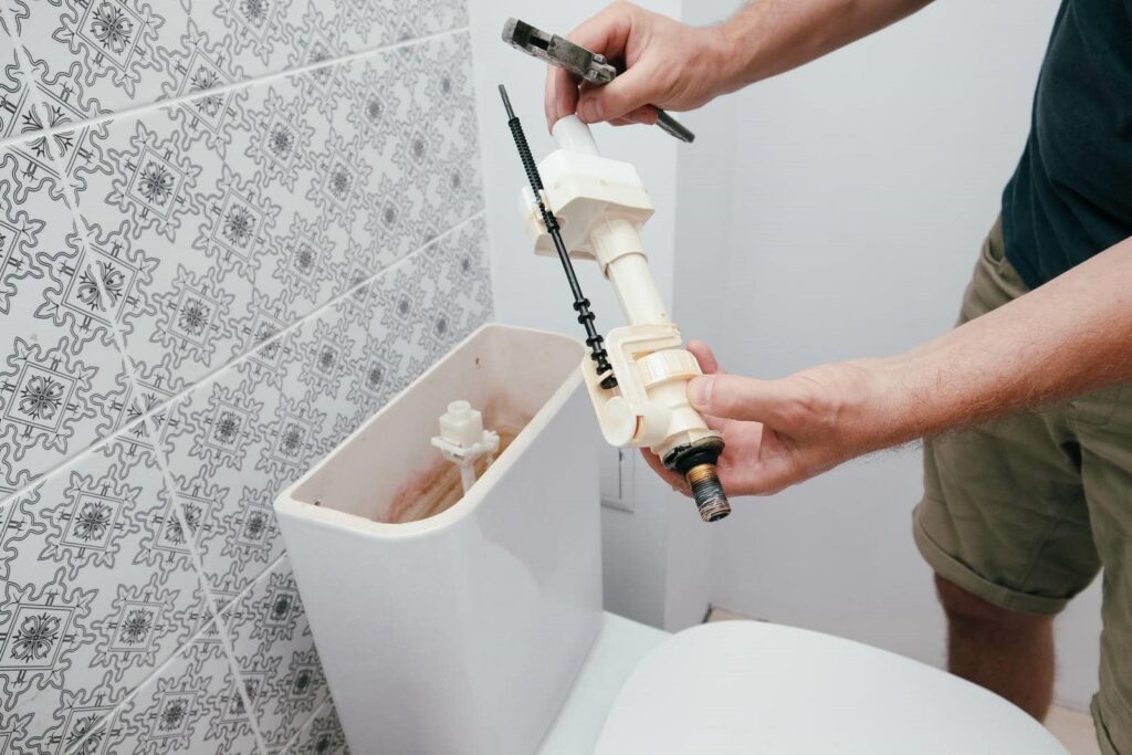 Hands holding a toilet fill valve above an open toilet tank with patterned wall tiles.